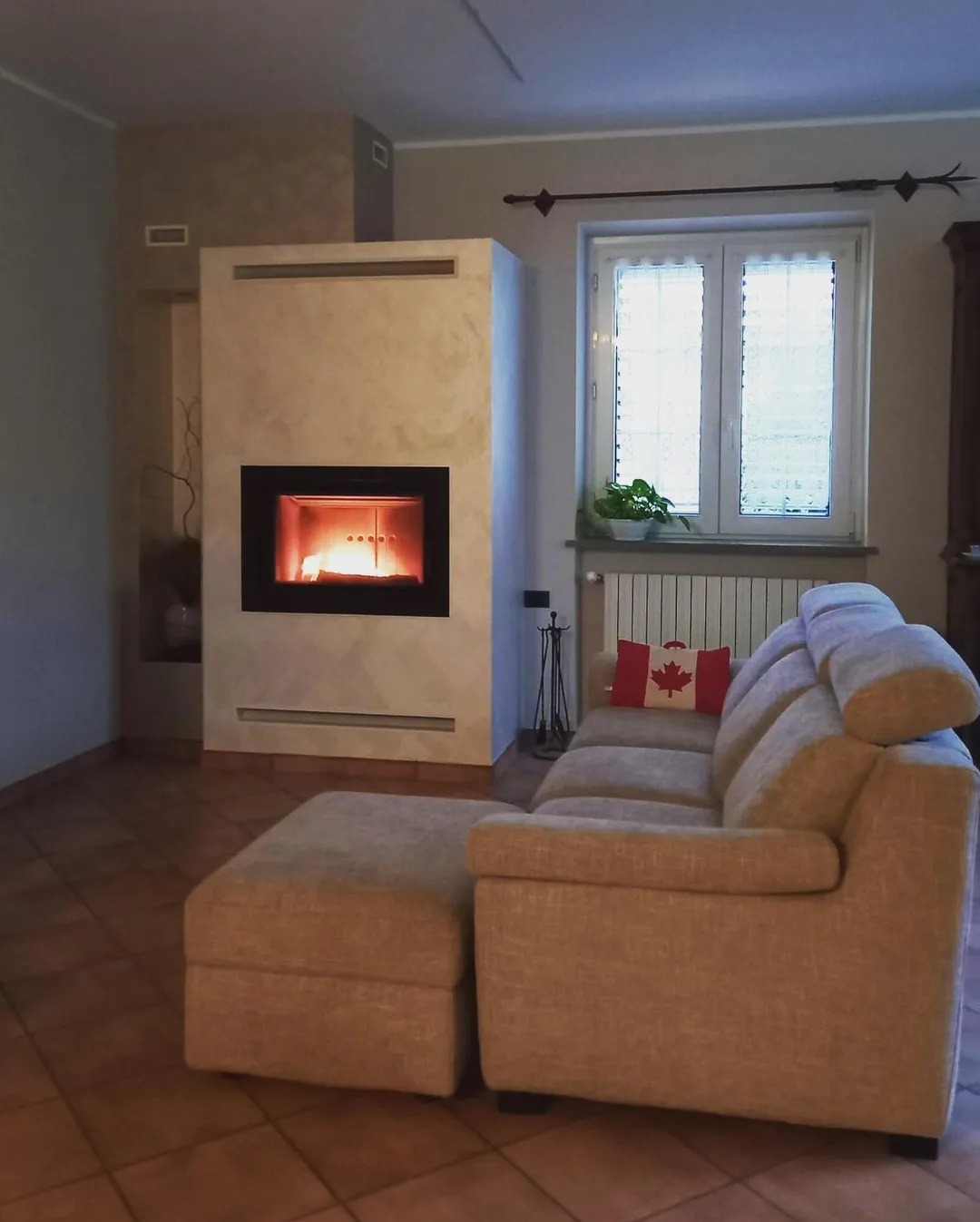 Nowoczesny, zabudowany w ścianie wkład z paleniskiem, obok beżowa sofa z poduszką w barwach Kanady, na tle okna z roletami.