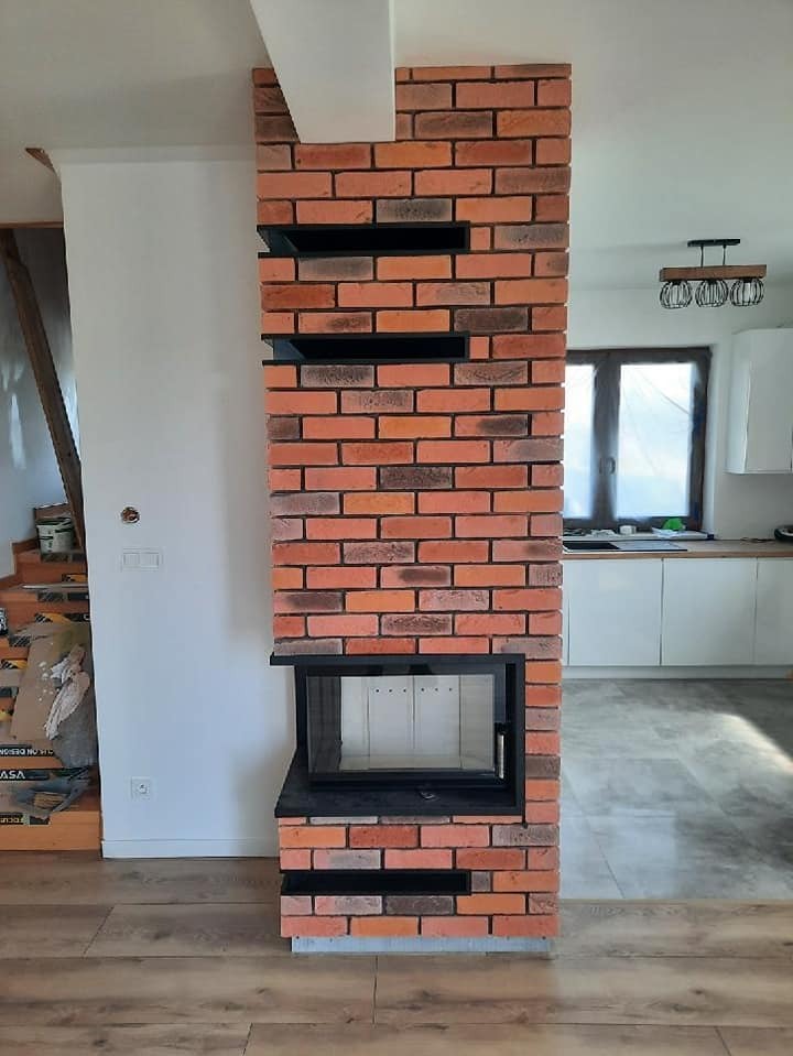 Kominek z cegły klinkierowej z czarną ramką i półkami w nowoczesnym salonie. Widoczne elementy wystroju wnętrza.