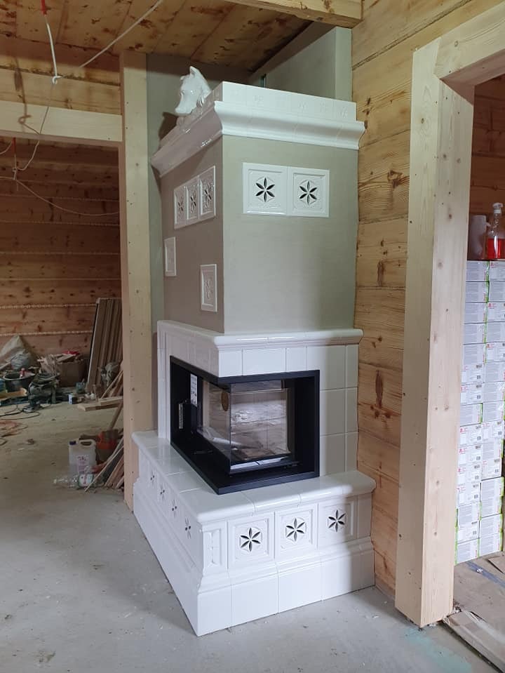 Biały, kaflowy kominek narożny z czarnym wkładem, zdobiony motywem kwiatowym, w surowym wnętrzu drewnianego domu. Widoczne materiały budowlane.