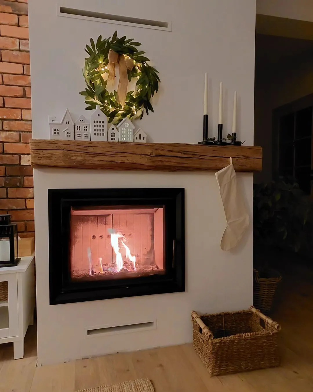 Ozdobiony kominek z palącym się ogniem, drewnianą półką, świątecznym wieńcem i dekoracjami w kształcie domków. Elementy cegły i kosze wiklinowe.