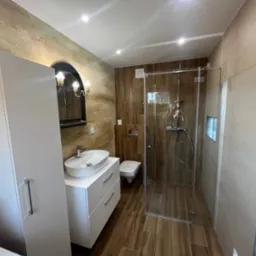 Nowoczesna łazienka z kabiną prysznicową ze szklanymi drzwiami, umywalką nablatową na białej szafce z szufladami i toaletą w tle, wykończona płytkami w odcieniach brązu i beżu, widoczny fragment...