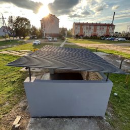 ProHome Sebastian Sulewski - Szara, pomalowana konstrukcja z ceglanym wnętrzem i metalowym dachem na zewnątrz, w tle budynki mieszkalne i zachodzące słońce.