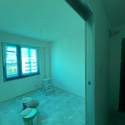 ProHome Sebastian Sulewski - Pokój w trakcie prac wykończeniowych: zabezpieczone okno folią, drabina, wiadra z farbą. Widoczne profile przy drzwiach. Ściany przygotowane do malowania.