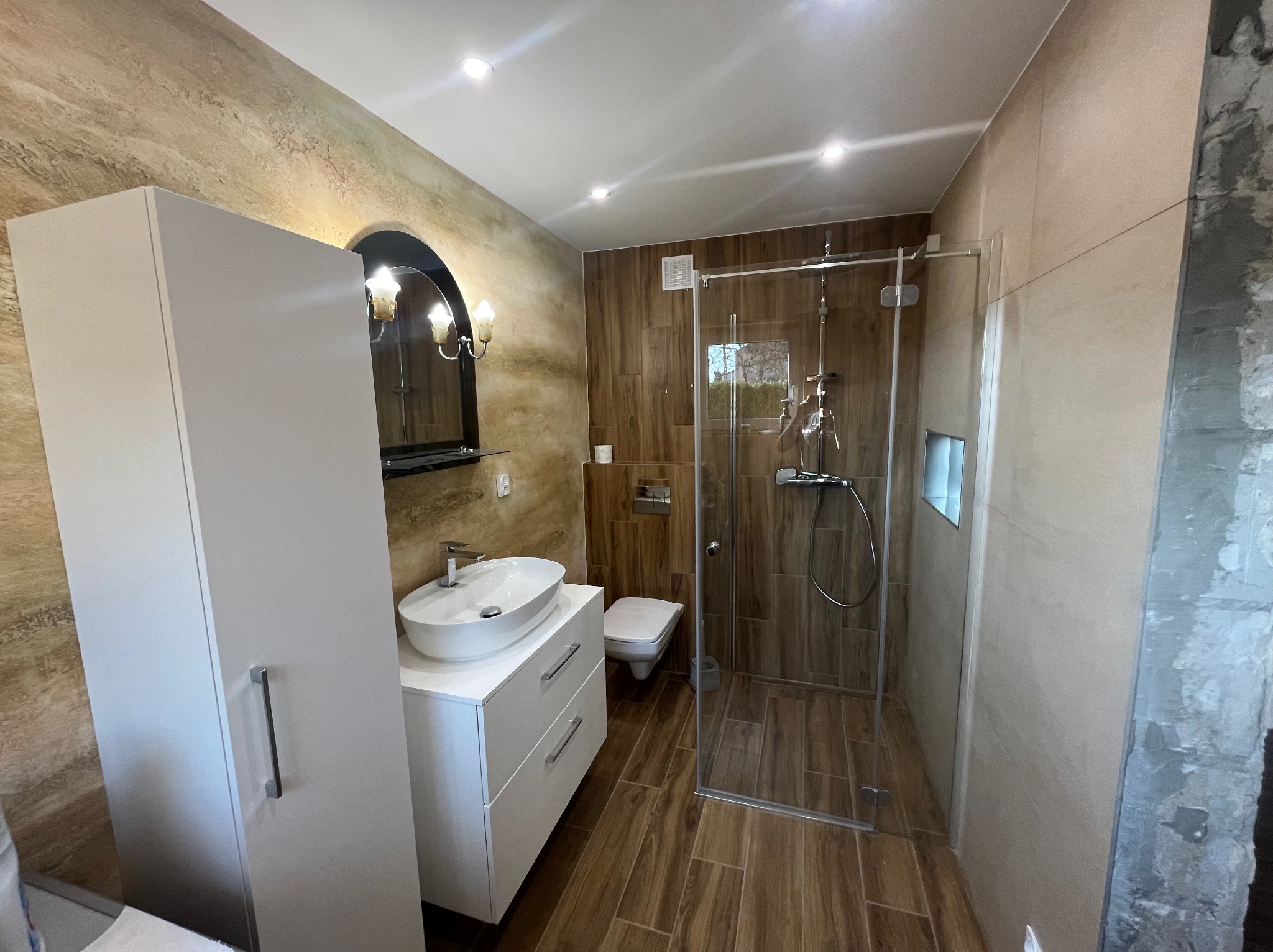 Nowoczesna łazienka z kabiną prysznicową ze szklanymi drzwiami, umywalką nablatową na białej szafce z szufladami i toaletą w tle, wykończona płytkami w odcieniach brązu i beżu, widoczny fragment...