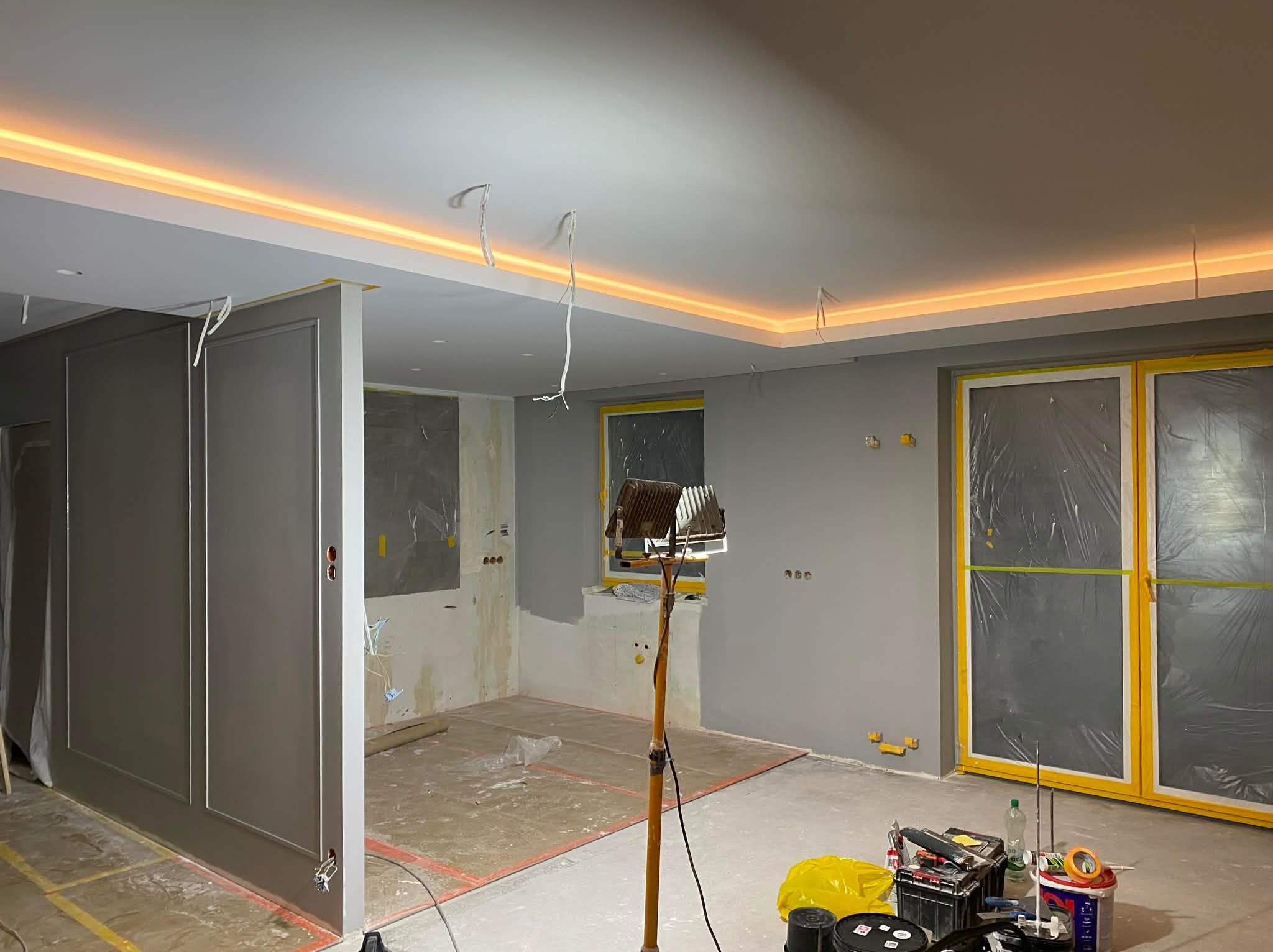 Wnętrze w trakcie prac wykończeniowych: szare ściany, zabezpieczone okna folią, widoczne narzędzia i oświetlenie halogenowe. Sufit z dekoracyjnym oświetleniem LED.