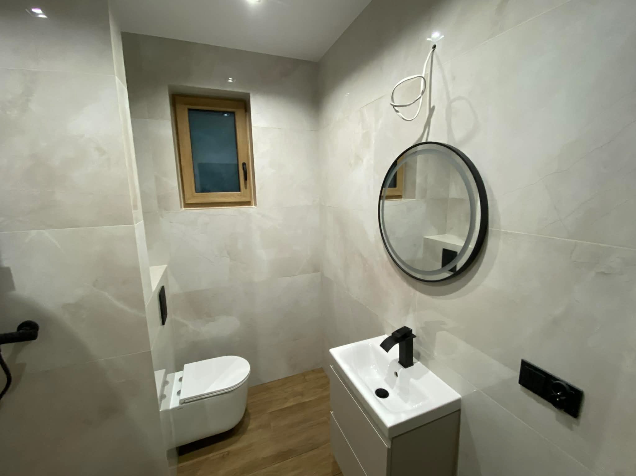 Nowoczesna łazienka po remoncie: jasne płytki, drewnopodobna podłoga, okrągłe lustro z czarną ramą, biała umywalka z czarną armaturą i podwieszana toaleta.