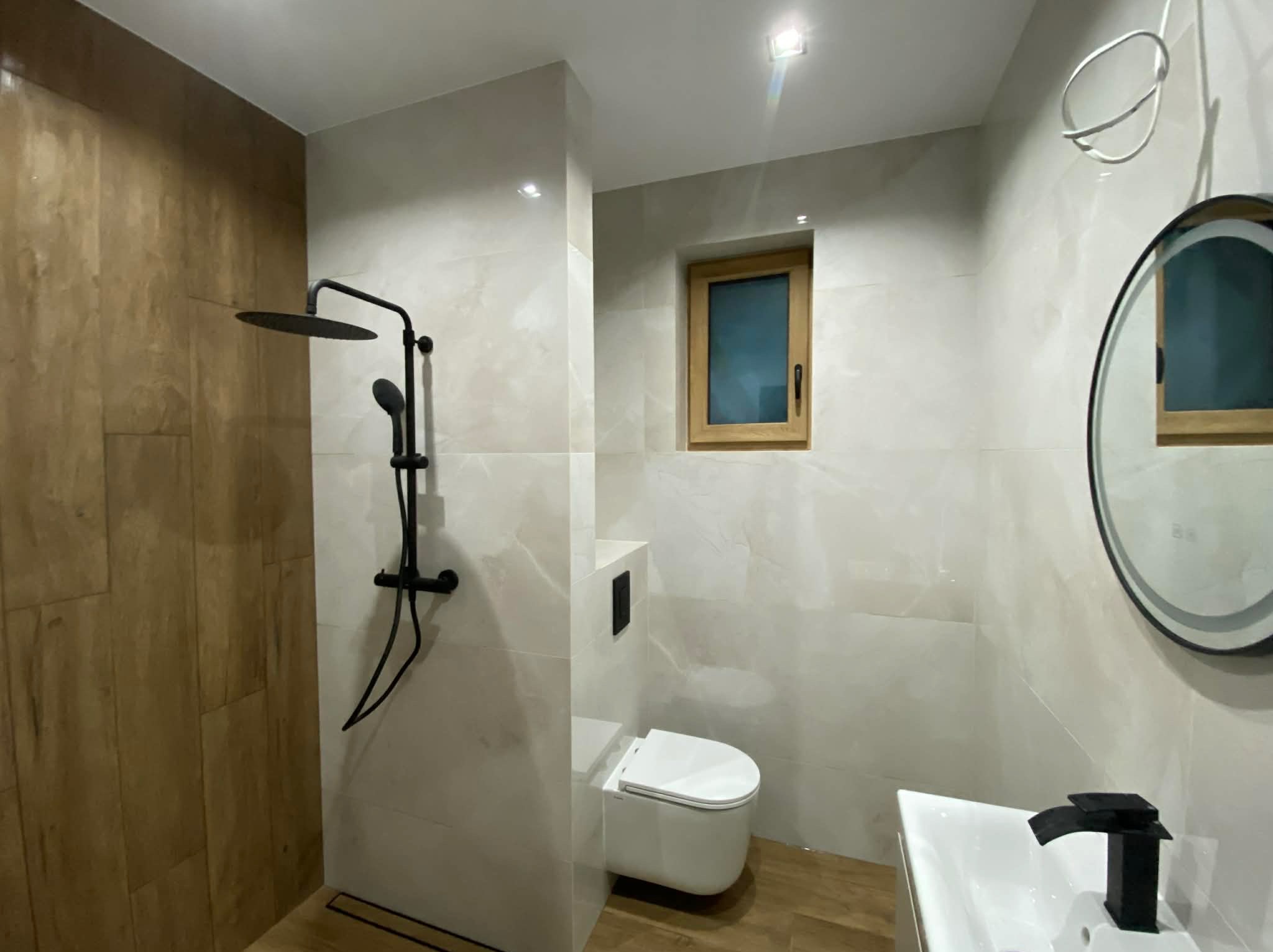 Nowoczesna łazienka z drewnopodobnymi płytkami, czarną armaturą i okrągłym lustrem z podświetleniem. Minimalistyczny design, jasne płytki imitujące marmur.