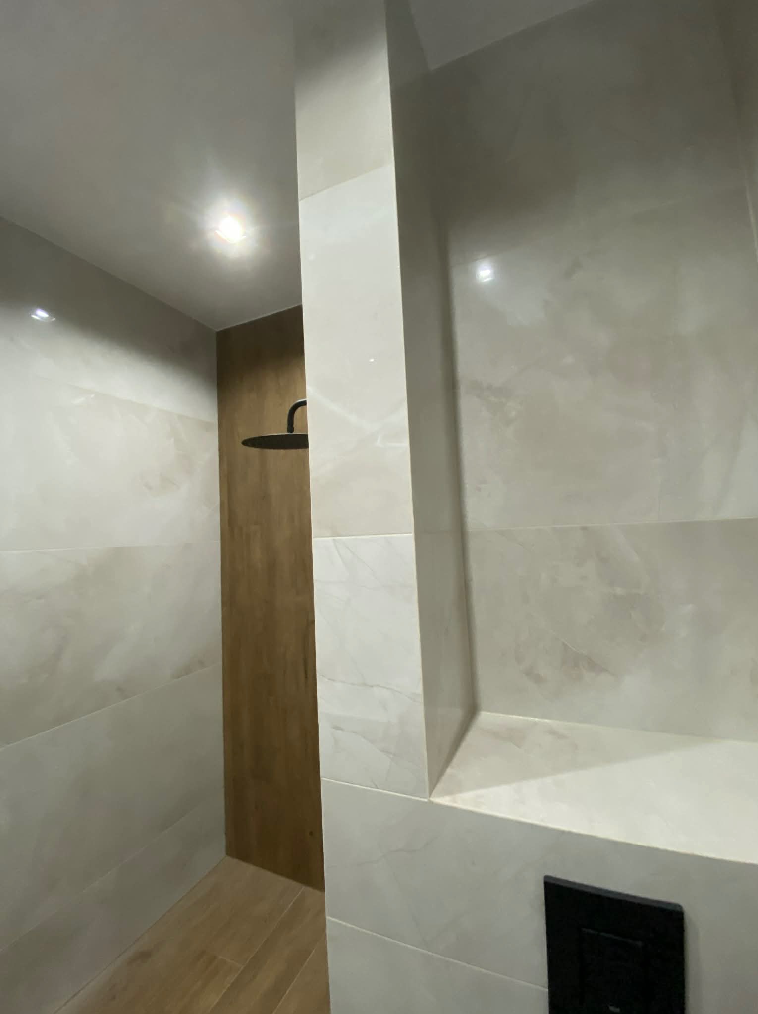 Wykończona kabina prysznicowa z beżowymi płytkami imitującymi marmur, drewnopodobną ścianą i czarną armaturą. Wnęka w ścianie wyłożona płytkami. Nowoczesny design.