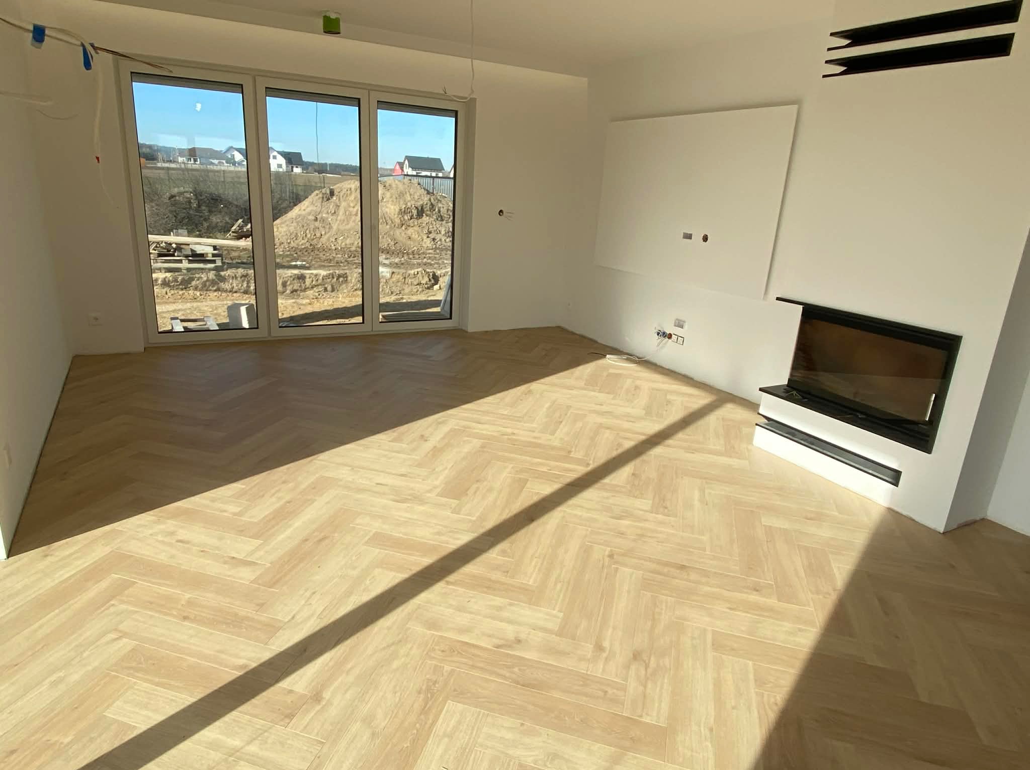 Jasny parkiet ułożony w jodełkę w przestronnym, nowoczesnym salonie z kominkiem i dużym oknem tarasowym. Widok na plac budowy za oknem.