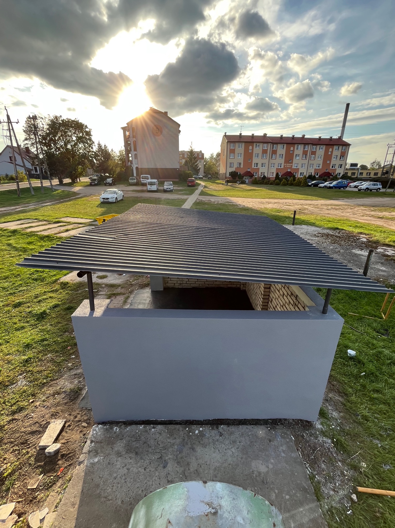 Szara, pomalowana konstrukcja z ceglanym wnętrzem i metalowym dachem na zewnątrz, w tle budynki mieszkalne i zachodzące słońce.