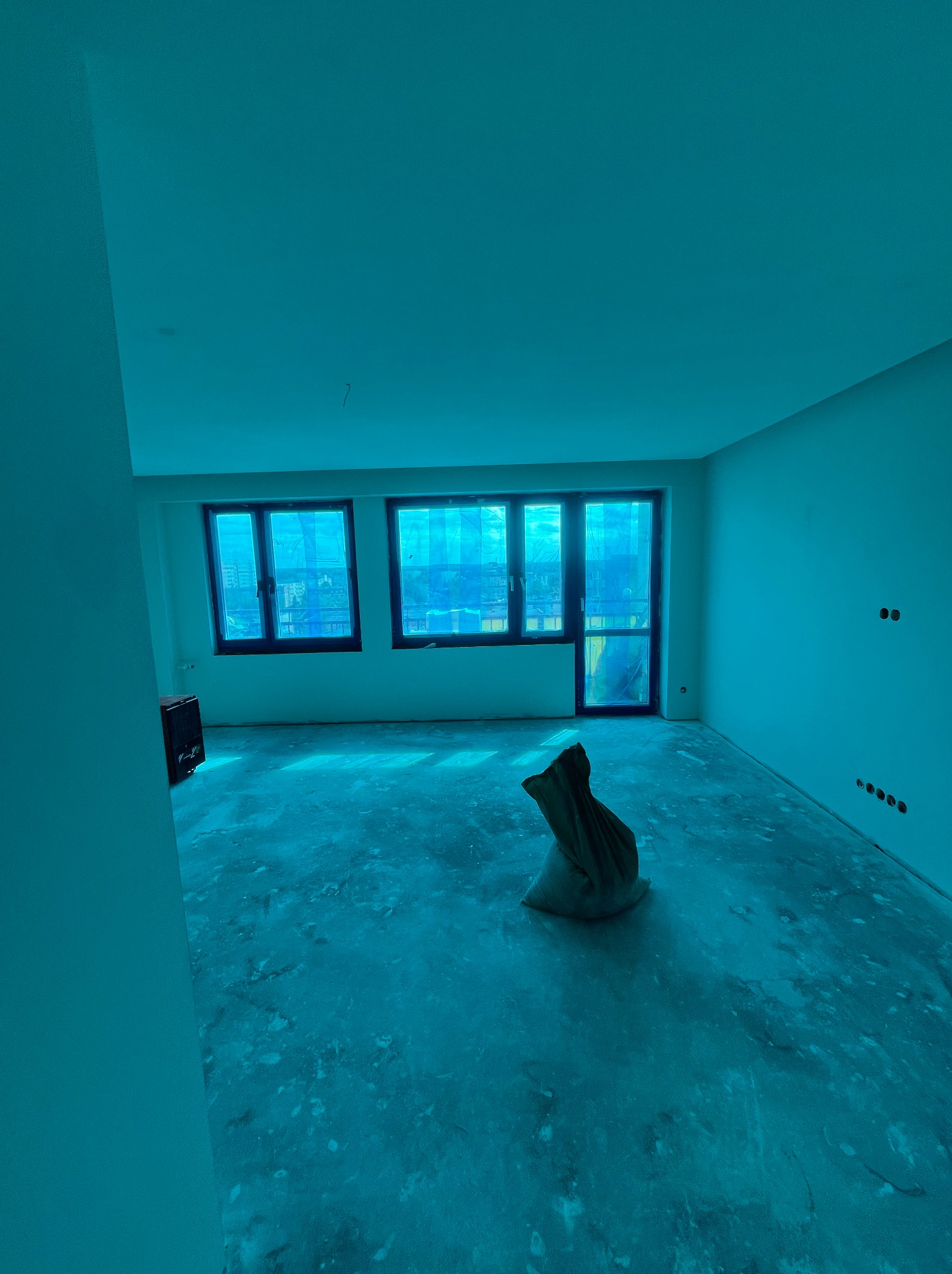 Wnętrze w trakcie prac wykończeniowych: surowe ściany, okna zabezpieczone folią, worek z materiałami budowlanymi na podłodze. Widoczne puszki elektryczne.