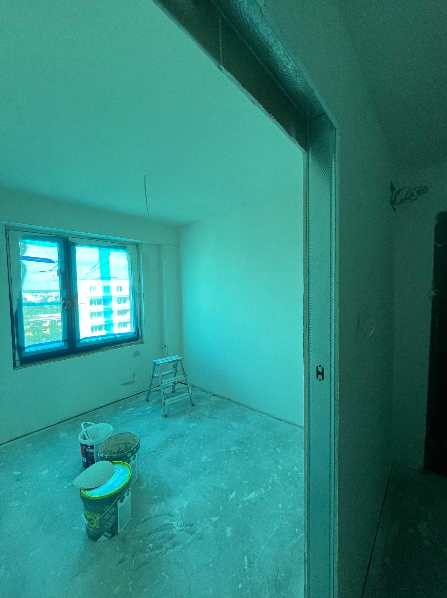 Pokój w trakcie prac wykończeniowych: zabezpieczone okno folią, drabina, wiadra z farbą. Widoczne profile przy drzwiach. Ściany przygotowane do malowania.