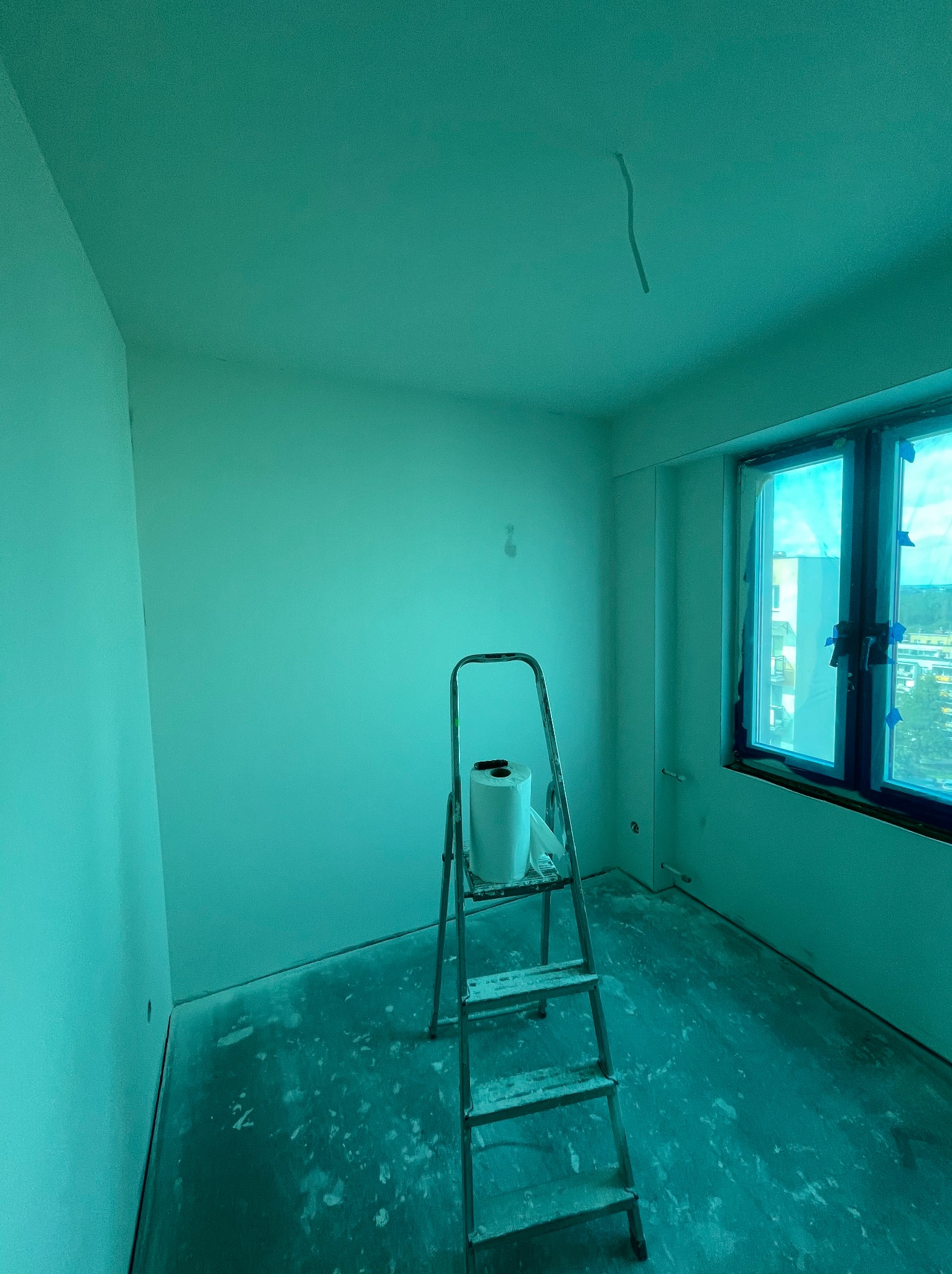 Pokój po świeżym malowaniu na zielono, z drabiną i rolką malarską. Okno zabezpieczone folią. Widoczne ślady farby na podłodze.