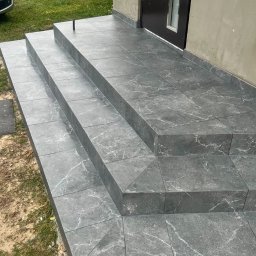 Bariev Andrzej - Schody wejściowe do domu wyłożone szarymi płytkami imitującymi marmur, z widocznym przycięciem płytek pod kątem na narożnikach i czarnym słupkiem na najwyższym stopniu.