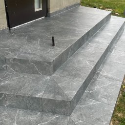 Bariev Andrzej - Zewnętrzne schody wejściowe wyłożone szarą płytką imitującą marmur, z wystającym prętem zbrojeniowym na górnym stopniu.