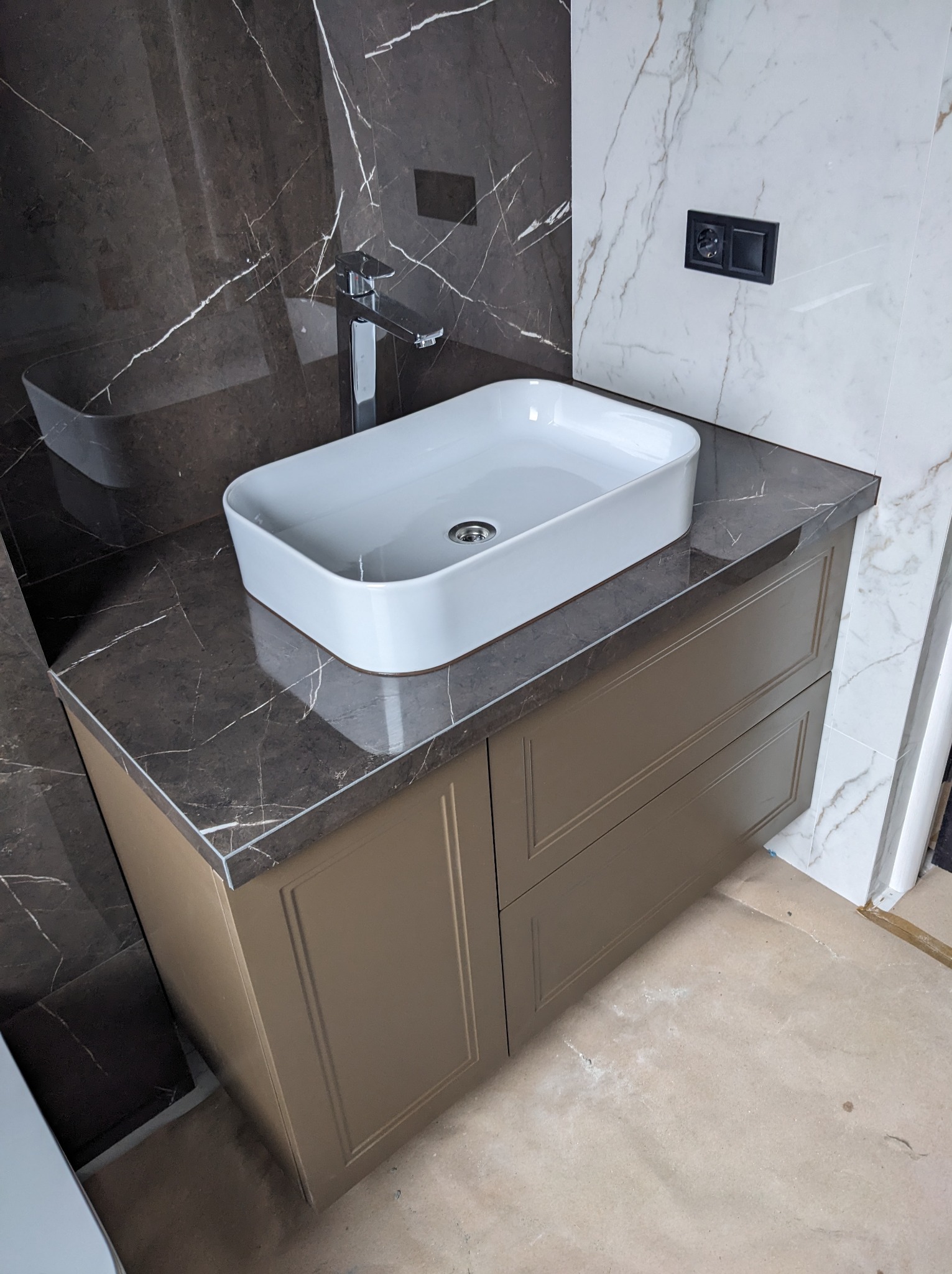 Nowoczesna łazienka z umywalką nablatową o prostokątnym kształcie, chromowaną armaturą i szafką w kolorze taupe z frezowanymi frontami, na tle płytek imitujących marmur w odcieniach brązu i bieli.