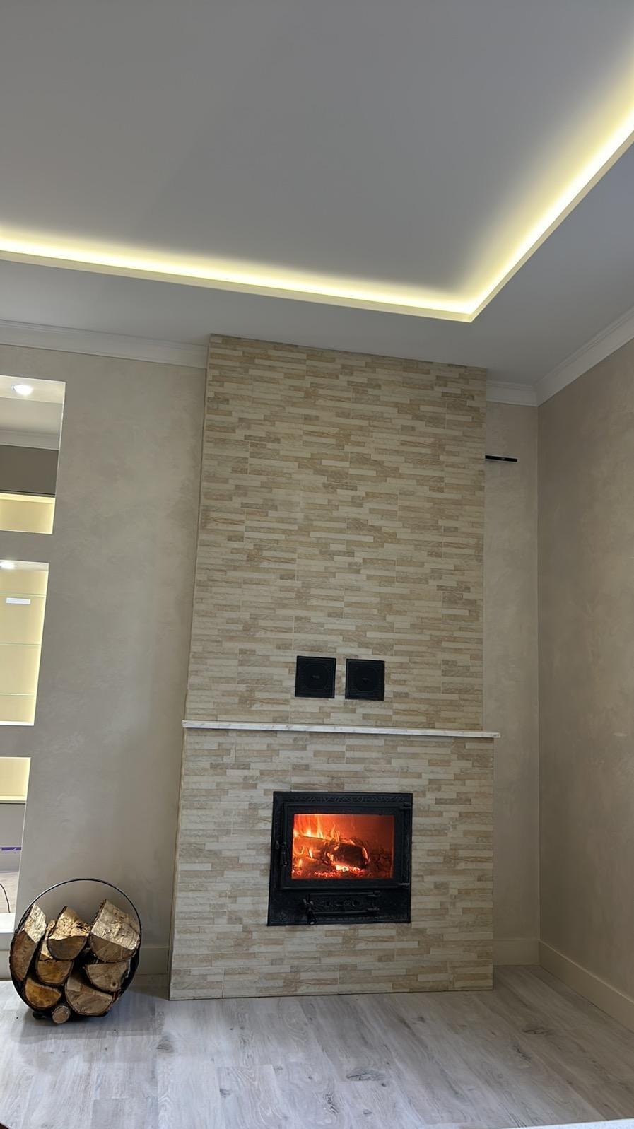 Kominek obudowany płytkami imitującymi kamień, z palącym się ogniem, drewno obok w metalowym koszu, podświetlany sufit.