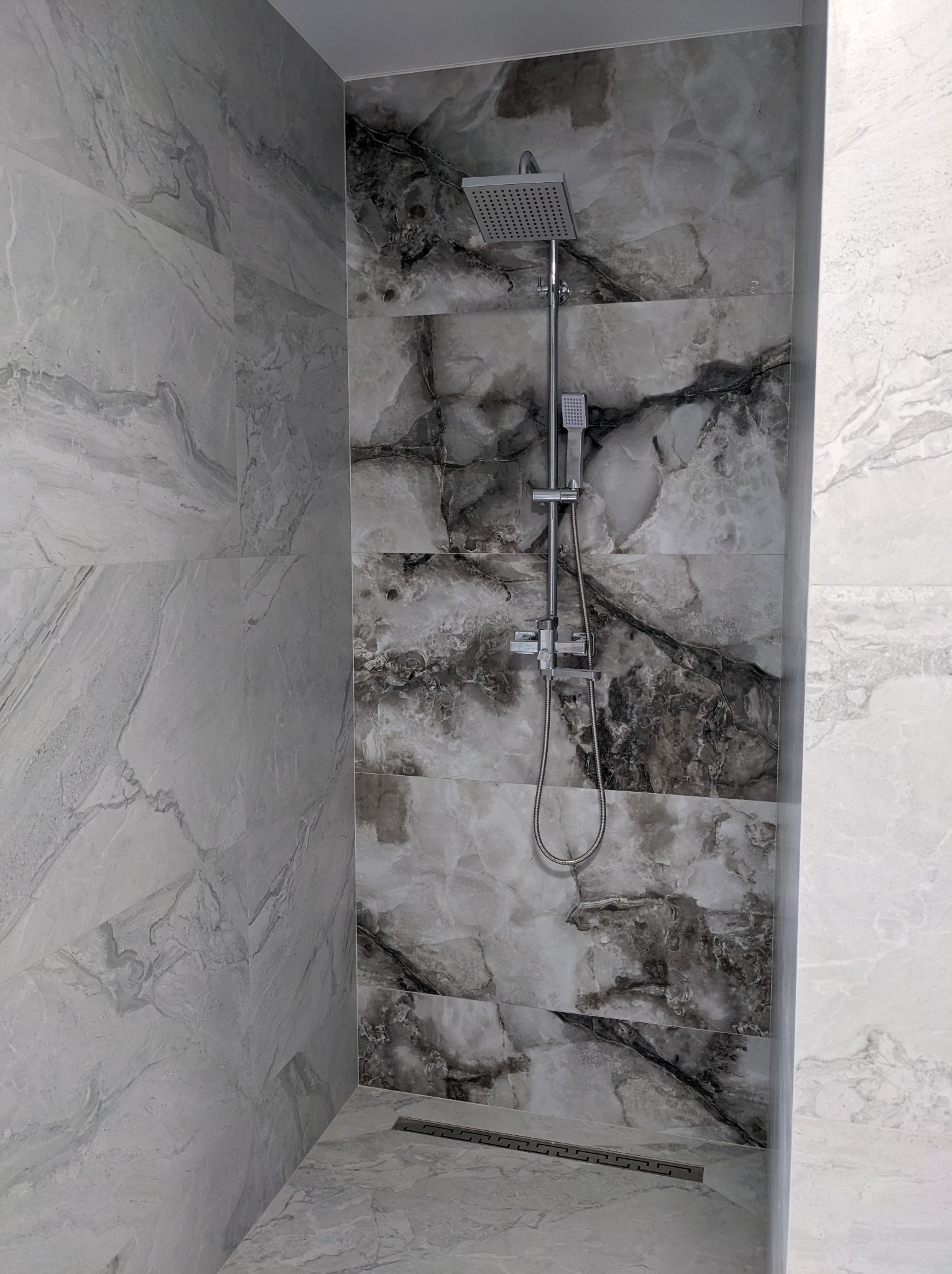 Kabina prysznicowa z deszczownicą, wykończona płytkami imitującymi marmur w odcieniach szarości i bieli, z odpływem liniowym w podłodze.