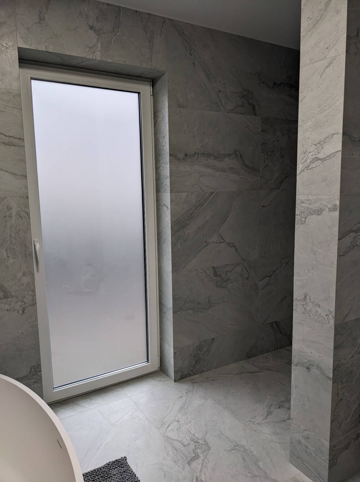 Nowoczesna łazienka wykończona płytkami imitującymi marmur, z oknem z matową szybą i fragmentem wolnostojącej wanny.