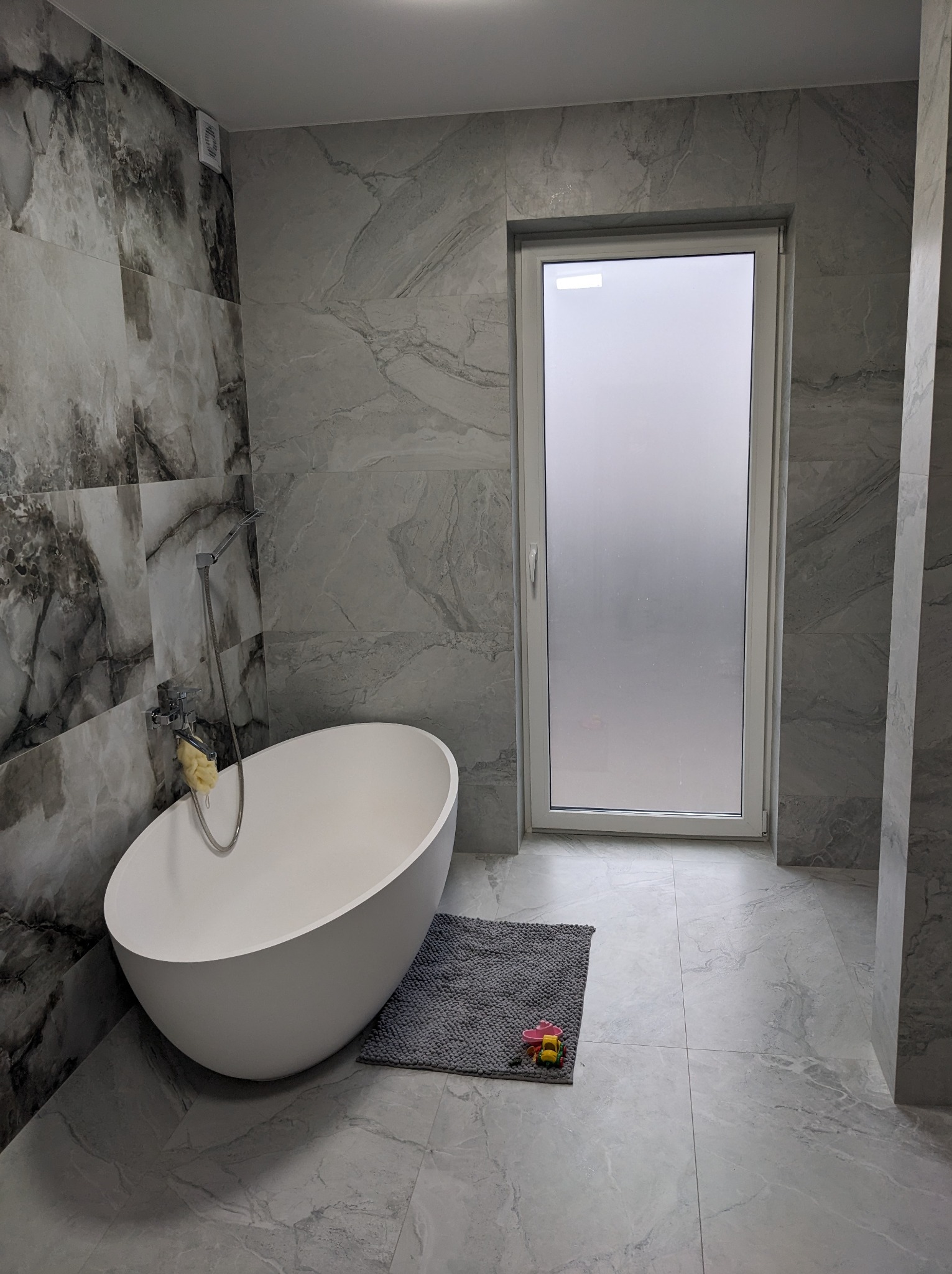 Nowoczesna łazienka z owalną białą wanną wolnostojącą, szarymi płytkami imitującymi marmur na ścianach i podłodze, oraz szarym dywanikiem z dziecięcymi zabawkami.