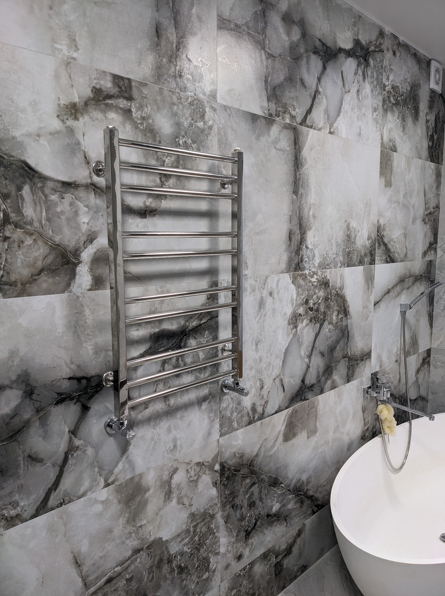 Wykończona łazienka z wanną wolnostojącą i grzejnikiem drabinkowym na ścianie wyłożonej płytkami imitującymi marmur w odcieniach szarości i bieli.
