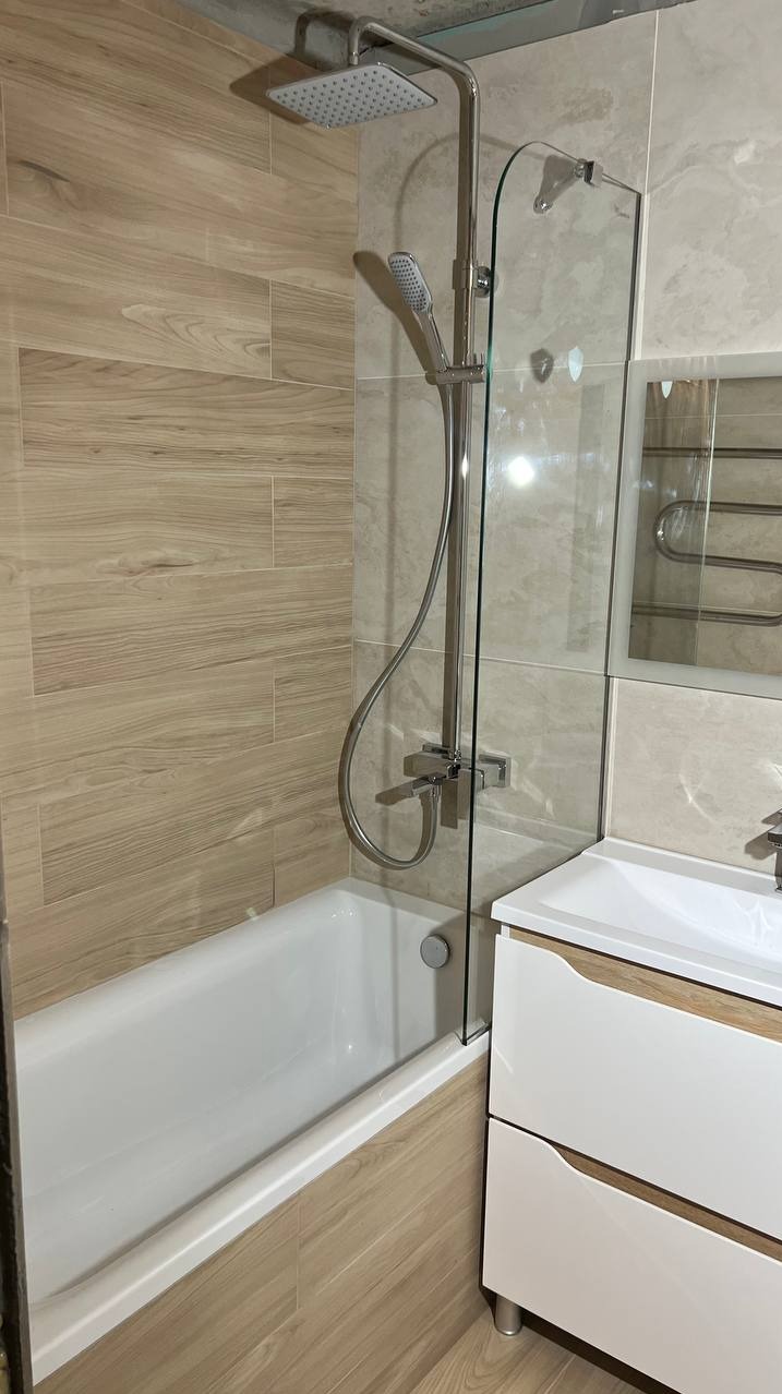 Wykończona łazienka z wanną obudowaną płytkami imitującymi drewno, szklanym parawanem nawannowym, baterią prysznicową z deszczownicą i umywalką z szafką w kolorze białym.