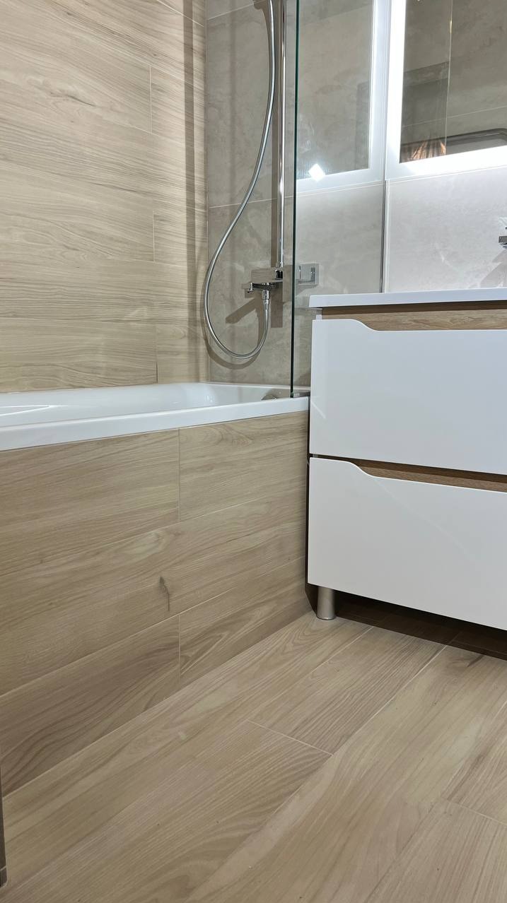 Nowoczesna łazienka z wanną obudowaną płytkami imitującymi drewno, białą szafką z drewnianymi akcentami i szklanym panelem prysznicowym.