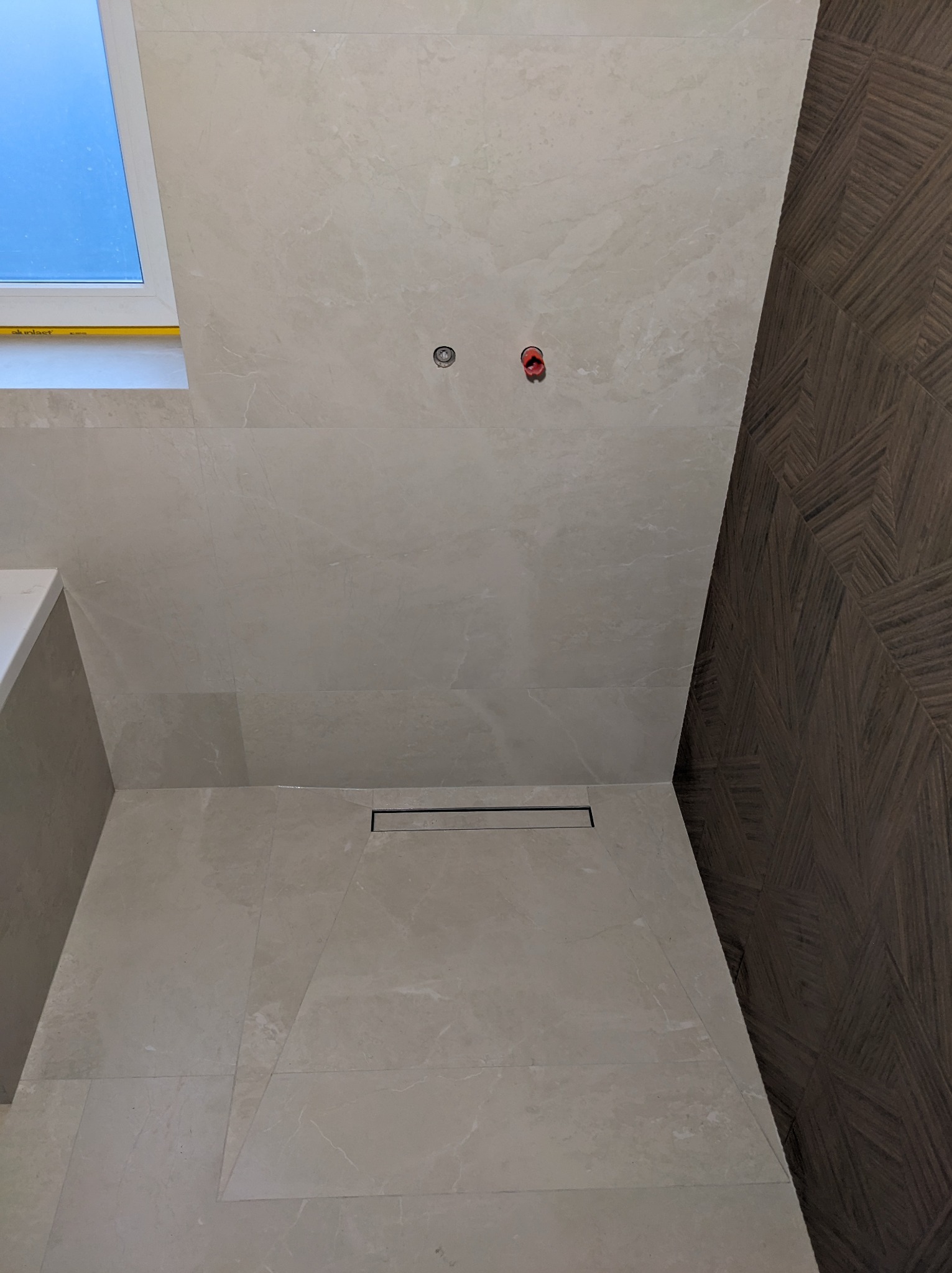 Beżowe płytki imitujące marmur na ścianie i podłodze łazienki, z odpływem liniowym i fragmentem drewnopodobnej ściany z geometrycznym wzorem oraz oknem dachowym.