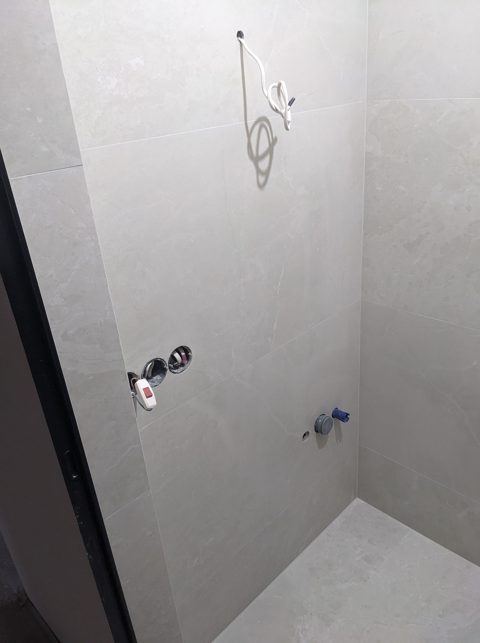 Ściana łazienki wyłożona jasnymi, marmurkowymi płytkami, widoczne otwory na armaturę i wystające kable elektryczne, z lewej strony fragment czarnej ramy drzwi.