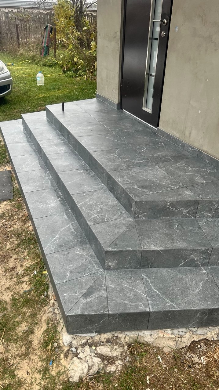 Schody wejściowe do domu wyłożone szarymi płytkami imitującymi marmur, z widocznym przycięciem płytek pod kątem na narożnikach i czarnym słupkiem na najwyższym stopniu.