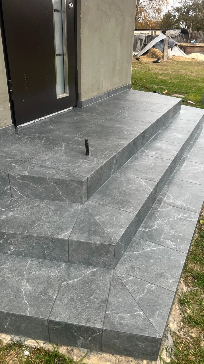 Zewnętrzne schody wejściowe wyłożone szarą płytką imitującą marmur, z wystającym prętem zbrojeniowym na górnym stopniu.