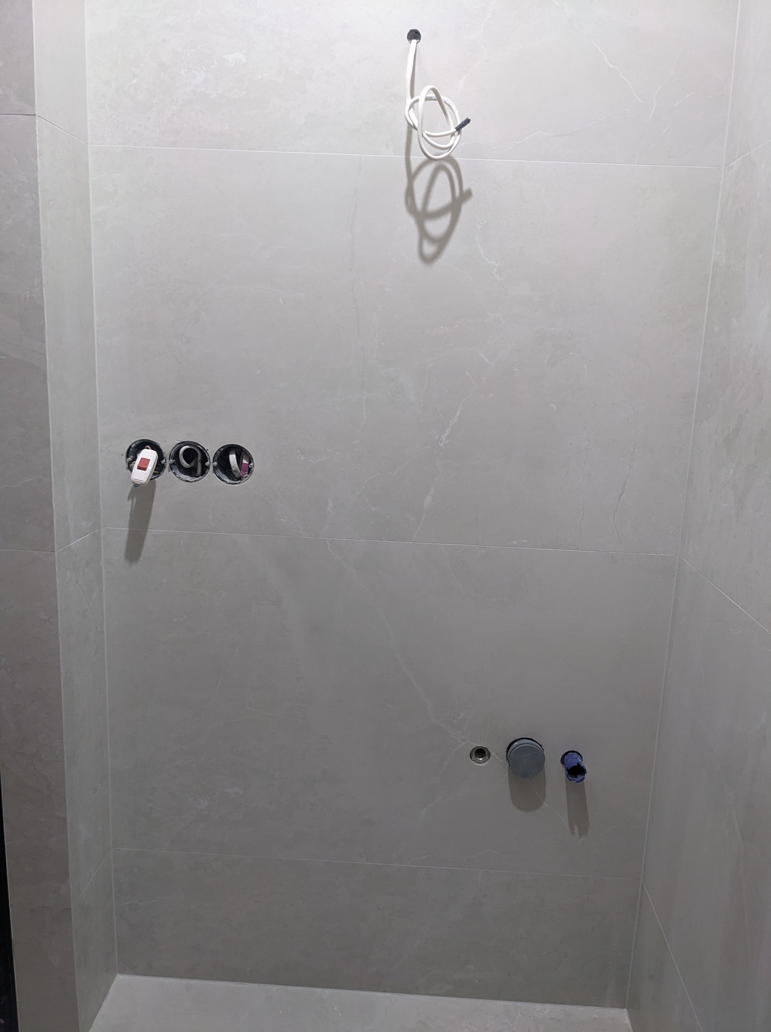 Ściana łazienki wyłożona szarymi płytkami, widoczne otwory na gniazdka elektryczne z wystającymi kablami, rury wodne i kabel oświetleniowy zwisający z sufitu, etap prac wykończeniowych.