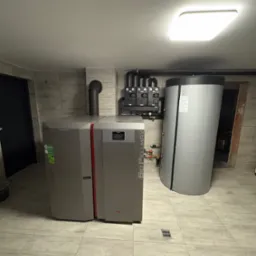 Szary piec na pellet z czerwonym akcentem i zbiornik akumulacyjny w kotłowni wyłożonej płytkami imitującymi drewno, widoczne czarne rury i armatura na ścianie.