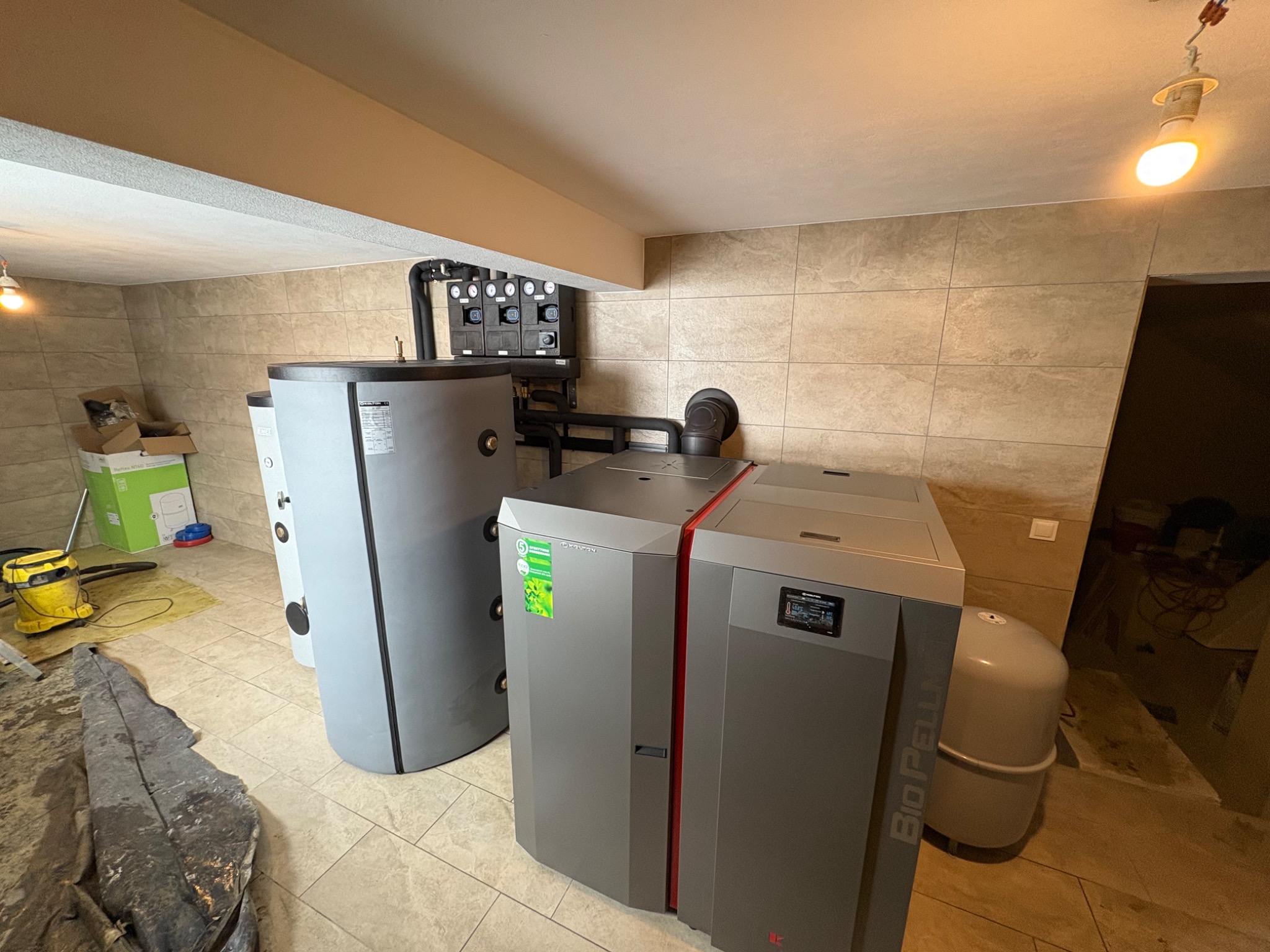 Szara kotłownia z piecem na pellet BioPell, zbiornikiem akumulacyjnym i osprzętem, widoczne manometry i rury, podłoga wyłożona płytkami, w tle kartony i odkurzacz.