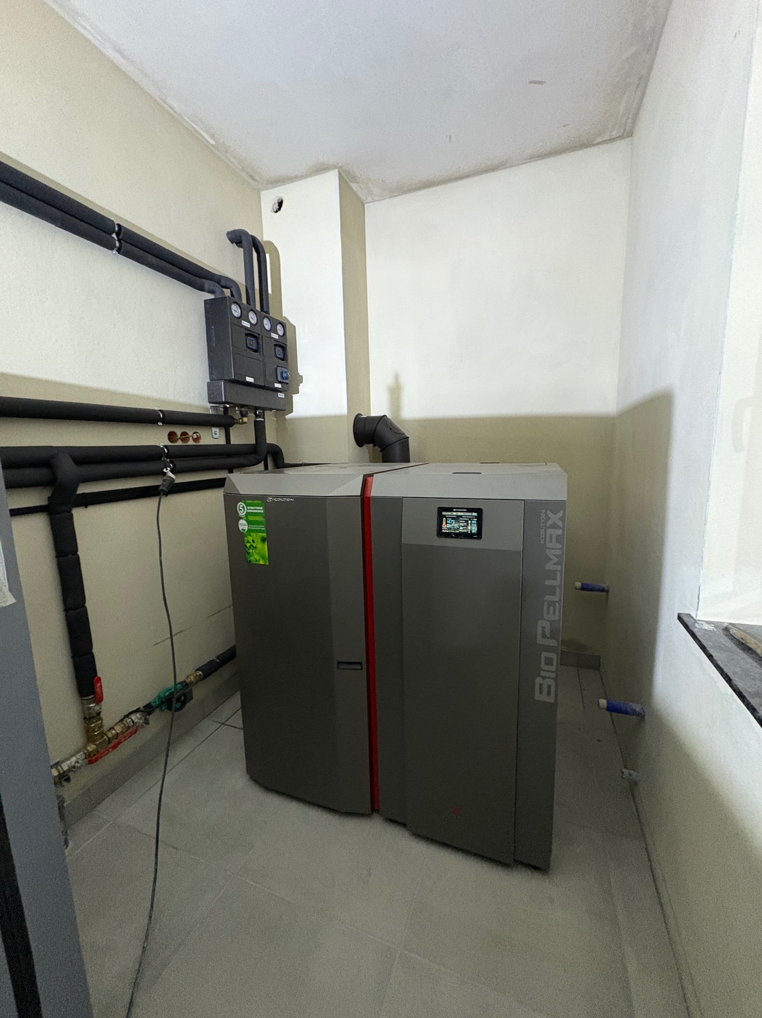 Szary piec na pellet Bio Pellmax z podłączonymi rurami, zainstalowany w pomieszczeniu z białymi ścianami i szarą podłogą.