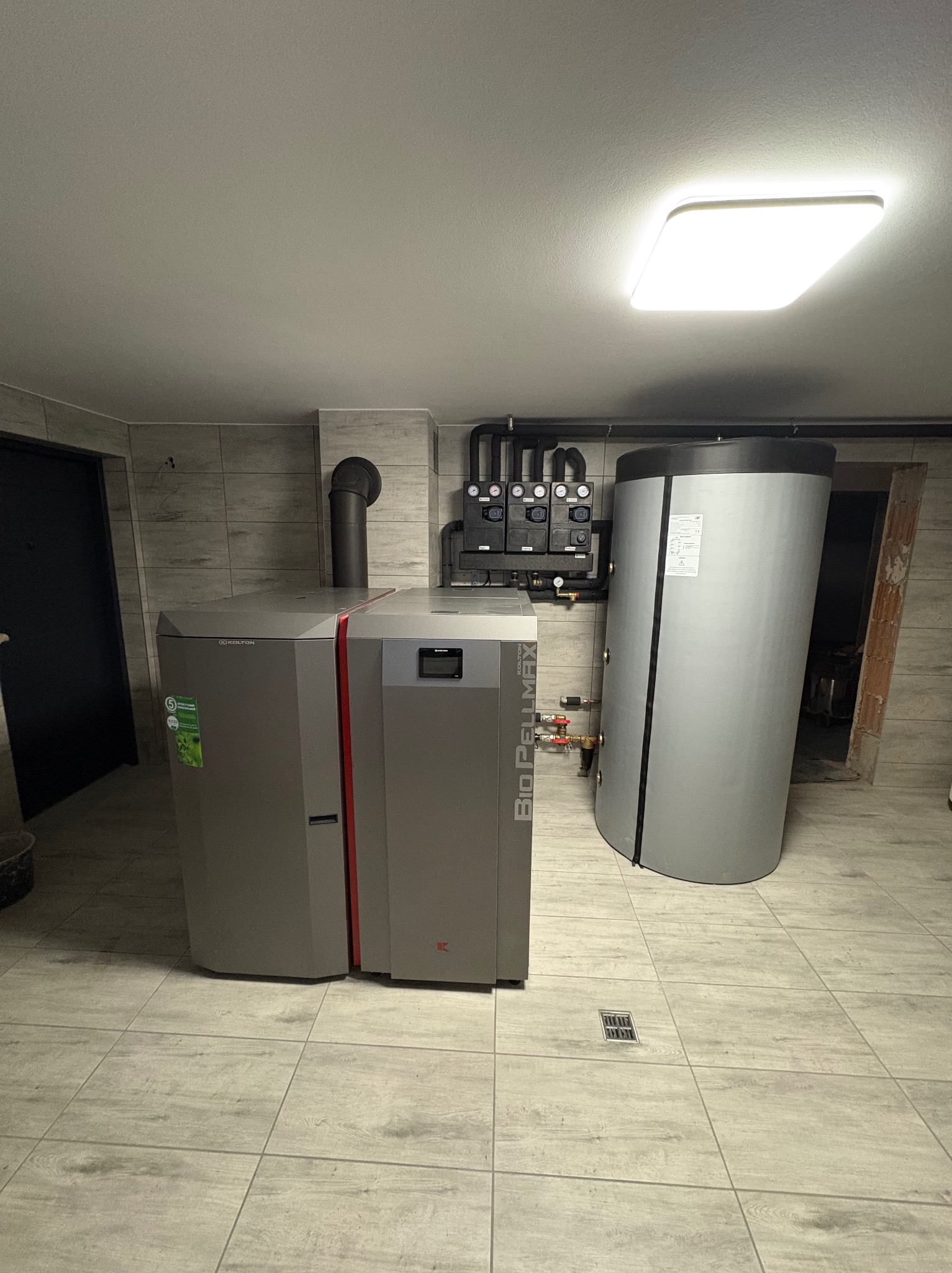 Szary piec na pellet z czerwonym akcentem i zbiornik akumulacyjny w kotłowni wyłożonej płytkami imitującymi drewno, widoczne czarne rury i armatura na ścianie.