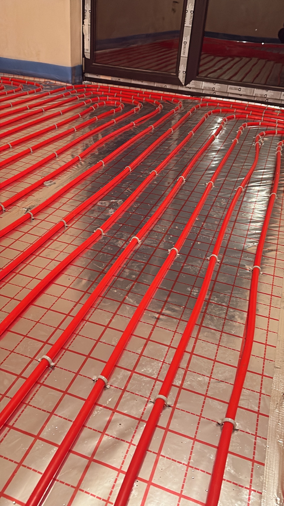 Czerwone rury ogrzewania podłogowego, równolegle ułożone na srebrnej folii z siatką, zamocowane białymi uchwytami, widok z góry, w tle okno.