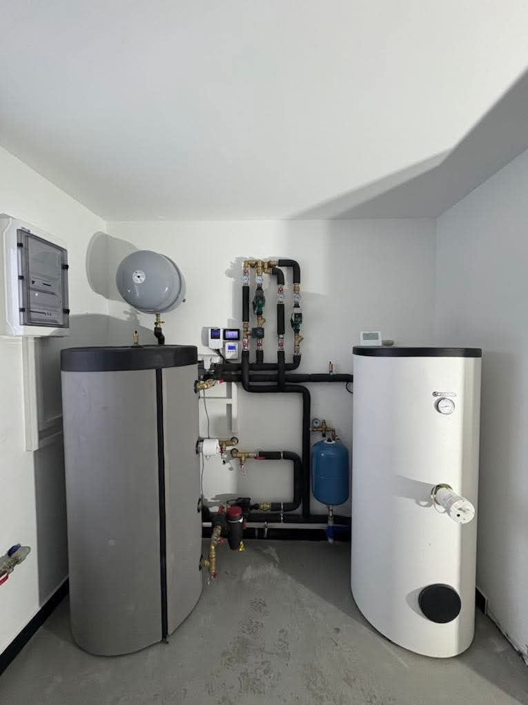 Dwa zasobniki ciepłej wody użytkowej w kotłowni, z widoczną instalacją hydrauliczną i elektryczną, szary zbiornik wyrównawczy pod sufitem, na szarym betonowym podłożu.