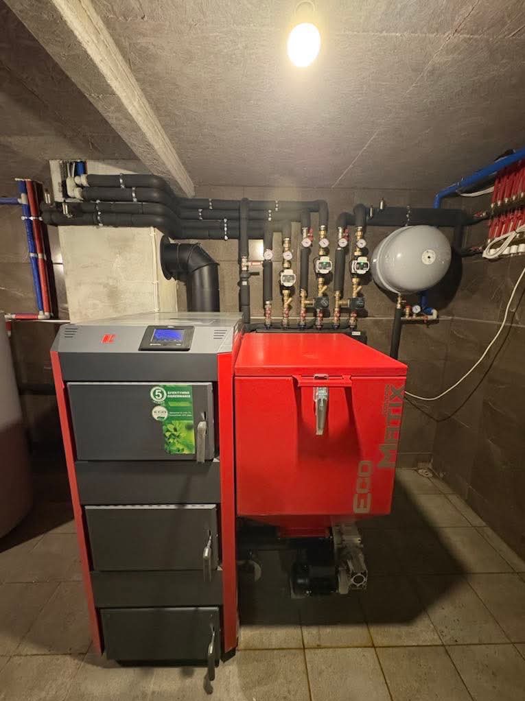 Nowoczesny piec CO z podajnikiem, szaro-czerwony, zainstalowany w kotłowni, widoczne liczne rury instalacji grzewczej pod sufitem, oświetlenie żarówką.