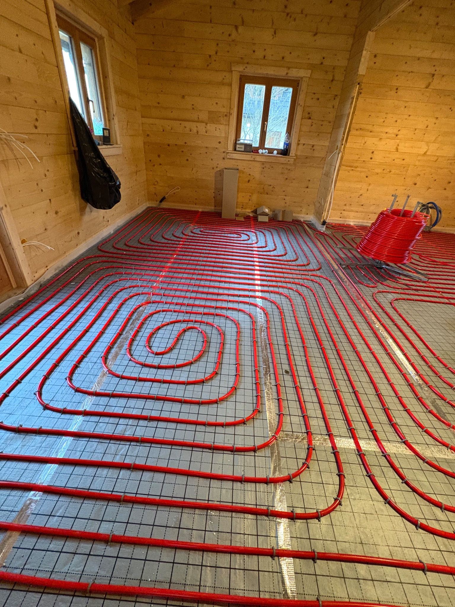 Czerwone rury ogrzewania podłogowego ułożone na folii w drewnianym domu. Widoczne okna i narzędzia. Instalacja przed zalaniem posadzką, etap prac.