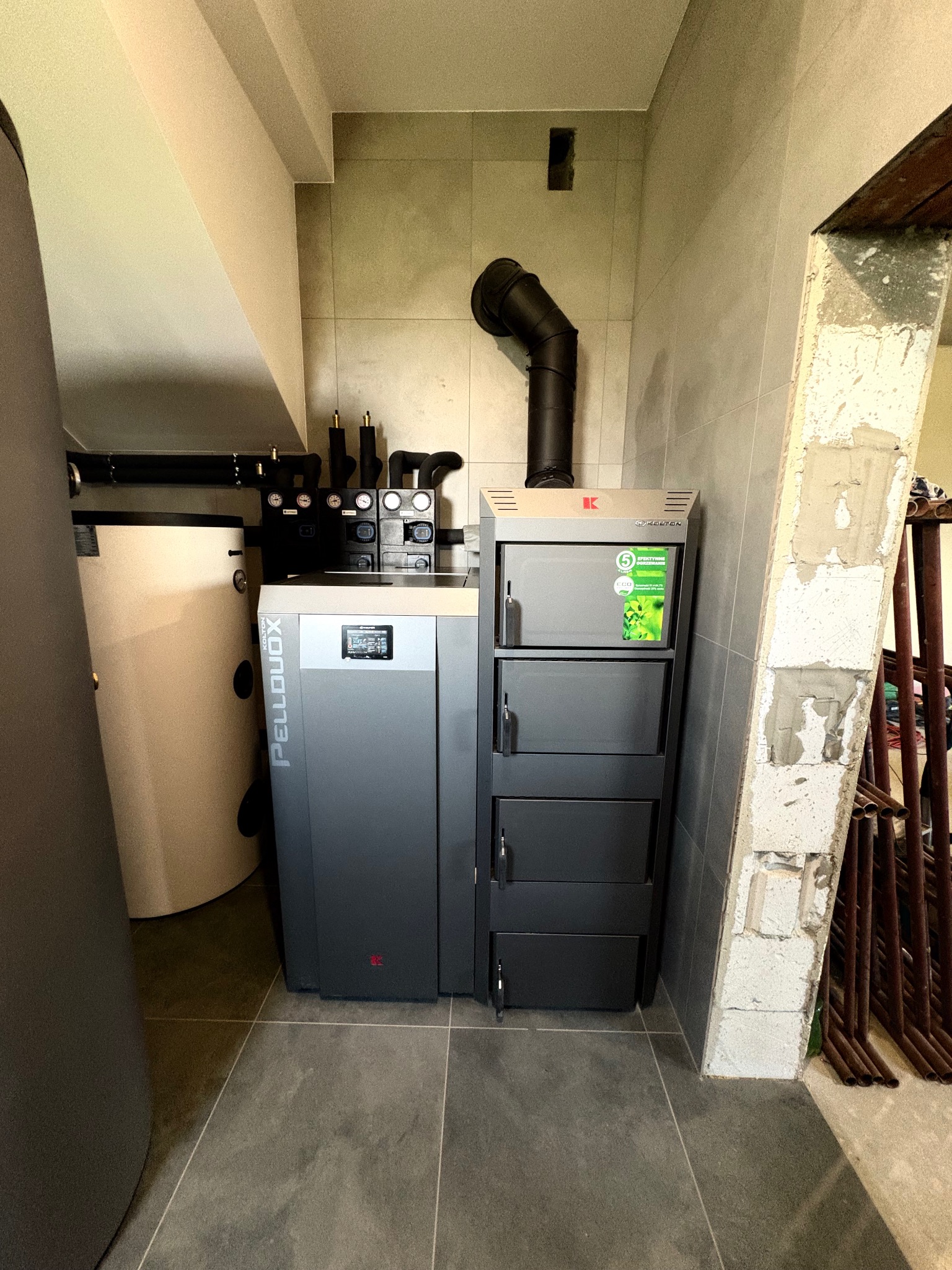 Szary piec na pellet z zasobnikiem, widoczny w kotłowni z szarą glazurą i elementami instalacji. Ujęcie z bliska, podkreślające nowoczesny design i funkcjonalność.