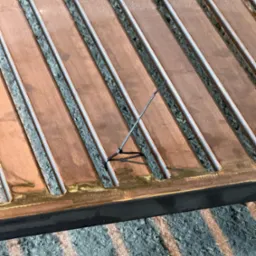 Metalowa konstrukcja z miedzianymi elementami wypełnionymi szarym kruszywem, zabezpieczona czarną opaską zaciskową.