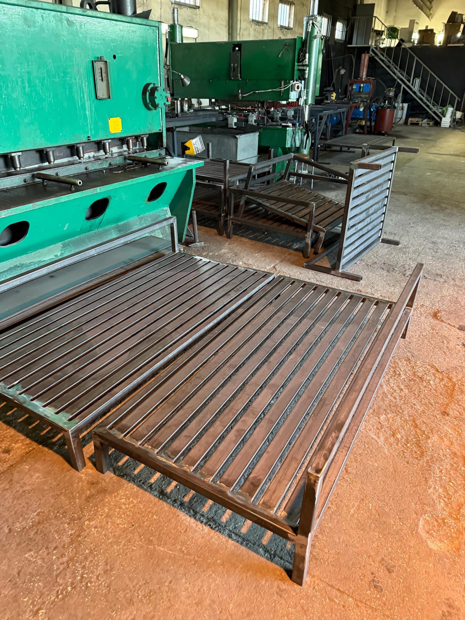 Metalowe konstrukcje ogrodzeniowe w warsztacie, widoczne surowe spawy i elementy przed obróbką wykończeniową, na pierwszym planie rama ogrodzenia z poziomymi prętami, w tle maszyna do cięcia metalu.