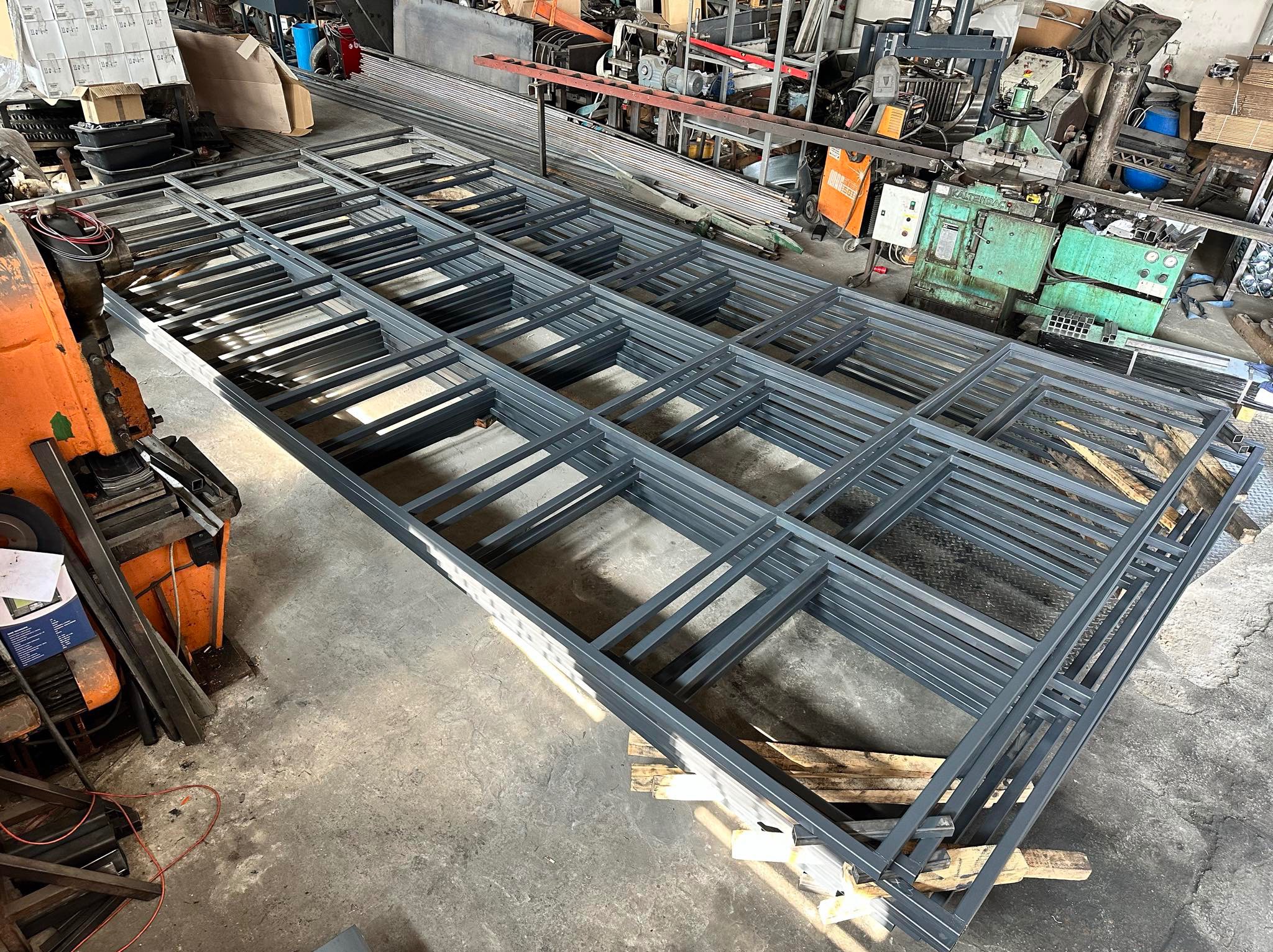 Stos ram metalowych, prawdopodobnie elementów ogrodzenia, pomalowanych na szaro, ułożonych na paletach w warsztacie z widocznymi maszynami do obróbki metalu w tle.