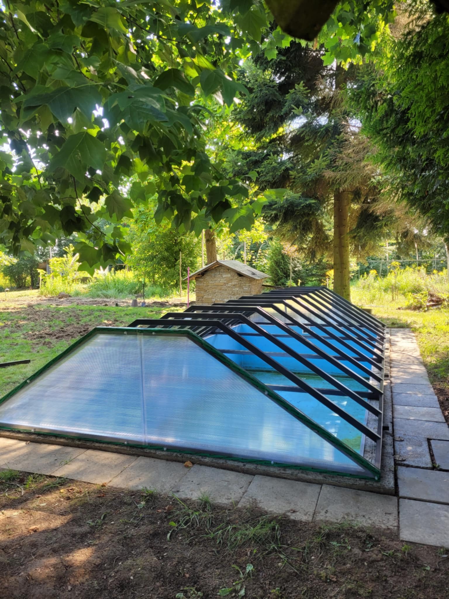 Zadaszenie basenu z poliwęglanu na betonowej podstawie, otoczone zielenią. Konstrukcja z czarnych profili, widok z niskiej perspektywy, elementy krajobrazu w tle.