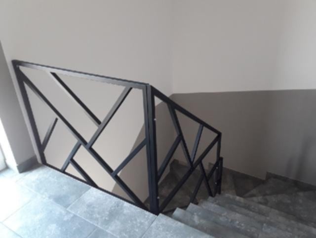 Nowoczesna, czarna balustrada metalowa o geometrycznym wzorze, zamontowana na schodach wewnątrz budynku. Szare płytki podłogowe i ściany w tle.