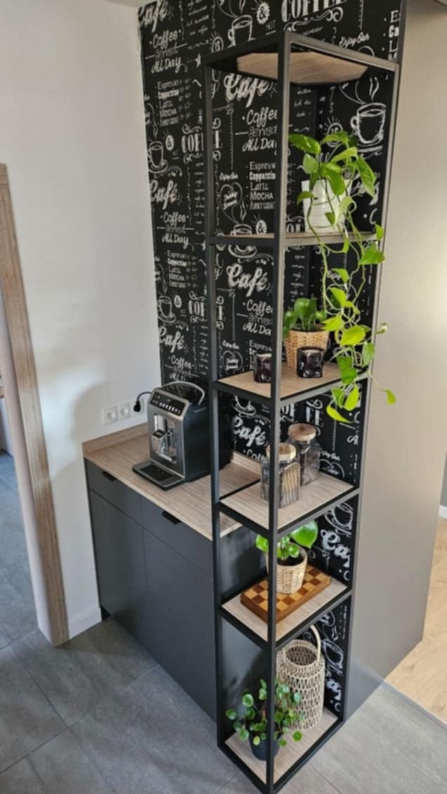Metalowy regał w stylu loft, z drewnianymi półkami, na których stoją rośliny, słoiki i szachownica. W tle ściana z motywem kawowym i ekspres do kawy.