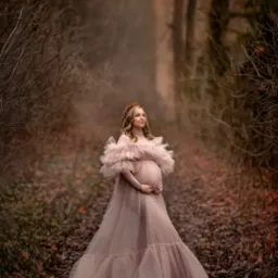 Kobieta w ciąży, w długiej, pudrowo-różowej sukni z falbanami, pozuje w lesie na tle ścieżki z opadłych liści, z koroną z brązowych kwiatów na głowie.