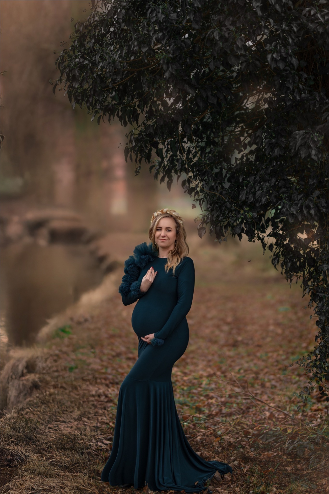 Kobieta w zaawansowanej ciąży, ubrana w dopasowaną, ciemnozieloną suknię z długim rękawem i ozdobnym, ciemnym kołnierzem z falban, pozuje na zewnątrz w naturalnym otoczeniu z drzewami i wodą w tle,...