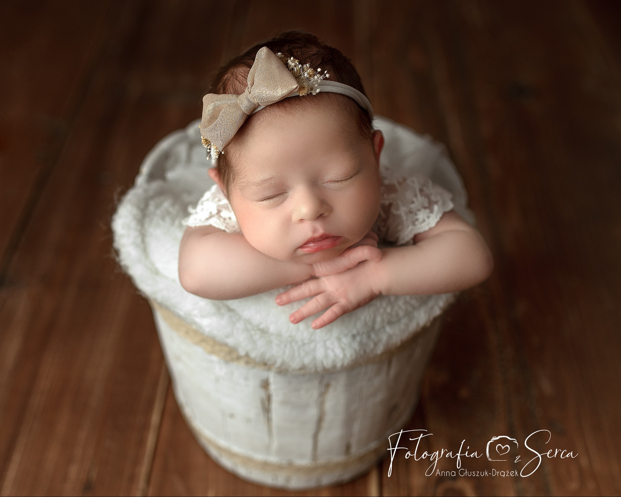 Śpiąca noworodka z beżową kokardą na głowie, oparta o brzeg białego, futrzanego wiaderka na tle drewnianej podłogi.